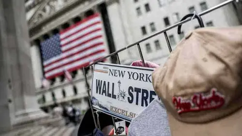 A Wall Street souvenir plate and New York cap on a street vendor’s rack, with the New York Stock Exchange and a large American flag in the background.
