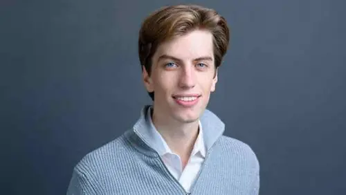 Leopold Aschenbrenner poses in front of a plain dark background, wearing a light gray zip-up sweater over a collared shirt.