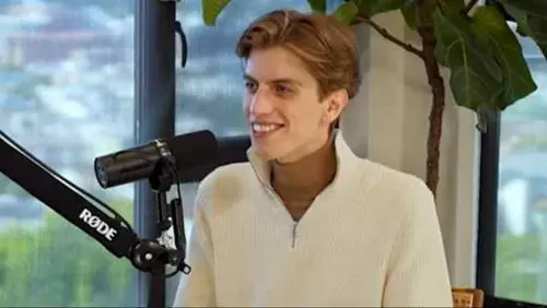 Leopold Aschenbrenner sits at a podcast microphone, smiling, wearing a light-colored zip sweater and brown turtleneck.