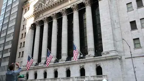 The front facade of the New York Stock Exchange with American flags and a person taking a photo in the foreground.